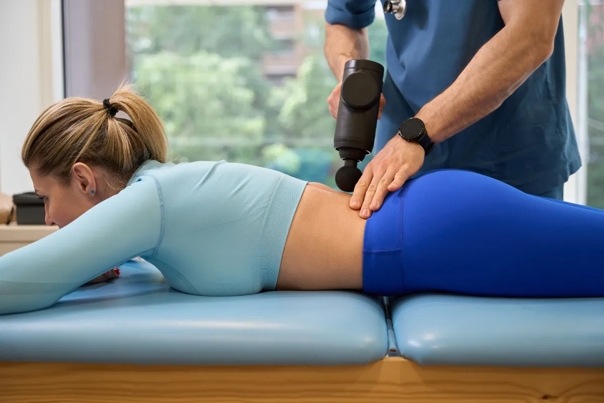 Mulher deitada em maca recebendo massagem na lombar feita por fisioterapeuta por meio de aparelho massageador
